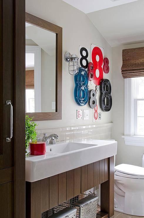 Beautifully decorated boy's bathroom features red, blue, and black number 8 art fixed over the toilet which is positioned beside a window dressed in a bamboo roman shade as a Duravit washstand boasts a shelf holding vintage metal locker bins and white porcelain sink located beneath a stained oak vanity mirror mounted on a light gray wall above porcelain accent tiles lit by vintage age wall sconces.