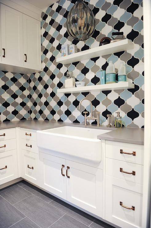 Gray blue and black Moroccan laundry room wall tiles create a stunning display surrounding white shaker cabinets with brass pulls and gray quartz countertops. The arabesque tiles liven up the space with a bold contrast displaying gray glass pendants and a set of stacked floating shelves stacked over a laundry farmhouse sink and gooseneck faucet.