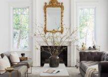 Elegant living room features a marble and brass coffee table placed on a gold and gray rug between side-by-side gray leather tufted accent chairs and a gray velvet chesterfield couch. An eye-catching gold baroque mirror hangs over a white fireplace mantel. A brass bar cart sits beside the fireplace.