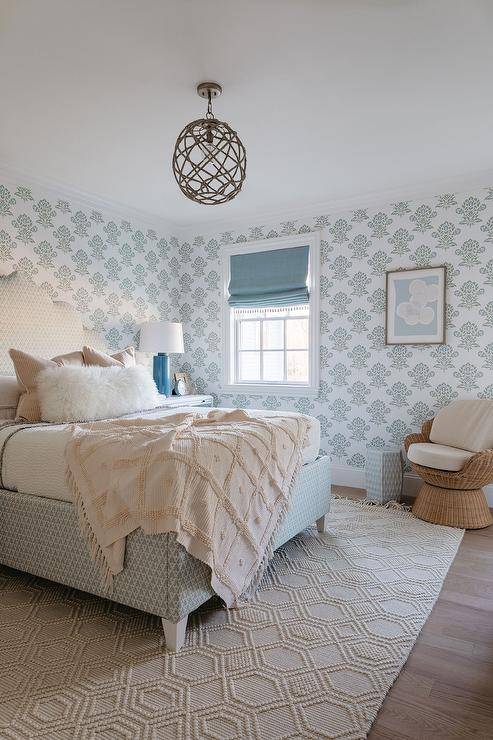 A macrame hexagon fringe rug is positioned beneath a white and blue Moroccan style bed topped with a white sheepskin pillow layered in front of pink stripe pillows complemented with an ivory macrame fringe throw blanket lit by a rope globe chandelier. Walls are clad in white and gray wallpaper frame windows dressed in a blue roman shade. A wicker swivel chair is placed beside a white bamboo stool table and beneath a blue and gray abstract art piece.