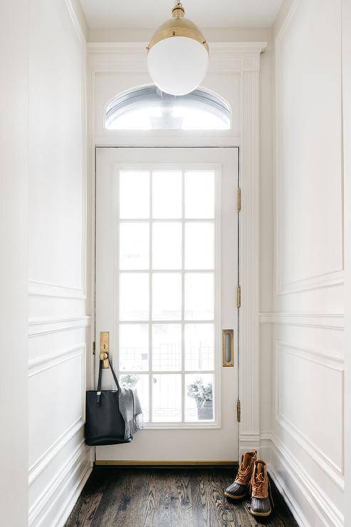 A glass paneled white door with a brass door knob and plate is located in a vestibule beneath an arched transom window. A brass Hicks pendant is hung between white walls with white trim and over a dark stained wood floor.