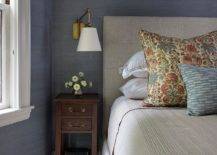 Bedroom features a light gray tufted bed on blue grasscloth wallpaper and a small tall wooden nightstand lit by a brass swingarm sconce.