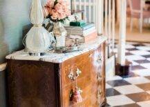 Black and white marble harlequin floor tiles bring a charming old-world finish to a foyer designed with a French dresser with pink tassels displaying a light blue glass lamp. The French dresser boasts styled decor against a staircase wall finished with green grasscloth wallpaper.