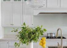 Hudson Valley Lighting Washington Pendant illuminates a white kitchen boasting off-white quartz countertops, white shaker cabinets, and polished nickel knobs. The custom kitchen island features a glass front beverage fridge along with a sink and polished nickel gooseneck faucet. Yellow accents deliver a cheerful, fresh appeal along with lush greenery.