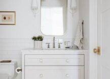 Aerin Fontaine Single Sconce flank a curved white mirror hung over an off-white dresser-like washstand finished with a polished nickel gooseneck faucet and white and gold half moon knobs. The washstand is mounted in front of a white subway backsplash tiles.