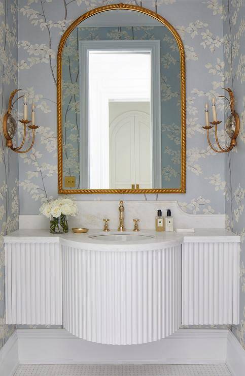 Romantic French-style bathroom is lit by facing gold candle sconces mounted on either side of a gold rope mirror hung from a wall covered in blue and white floral wallpaper. The mirror is mounted above a white half-moon French style sink vanity paired with a polished brass hook and spout faucet.