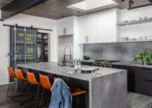 Stylish kitchen features a black center island complemented with a concrete waterfall edge countertop finished with a sink and a matte black pull out faucet. The faucet is mounted in front of brown molded leather counter stools illuminated by natural light from a skylight.