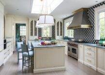 Hung beneath a skylight, white and gold pleated shade lights are positioned over a cream kitchen island accented with a gray marble countertop seating blue bamboo stools facing a sink with a brass faucet.