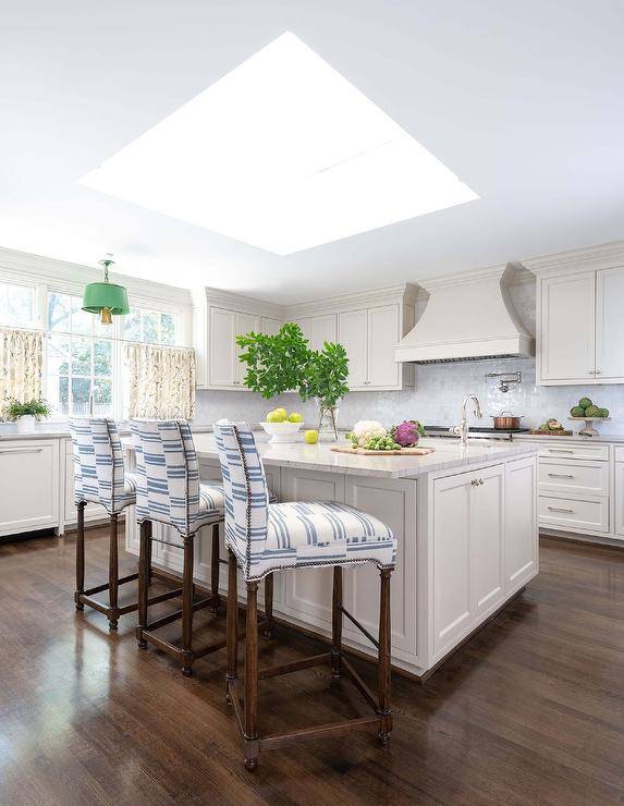 White and blue counter stools sit on a brown wood floor at a marble top island finished with a prep sink and a polished nickel gooseneck faucet lit by natural light from a large skylight.