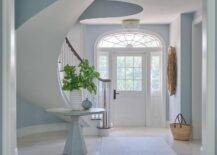 Morgan Harrison Home - A white front door with sidelights fixed beneath an arched transom window opens to a white and blue entry lit by an Aerin Jacqueline Flush Mount. A round oak table topped with white and gray vases and placed beside a curved staircase boasting a brown wood handrail on white spindles.