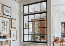 A taupe leather pillow top x-stool sits on a wood floor beneath interior windows, as walls are lined with dentil crown molding.