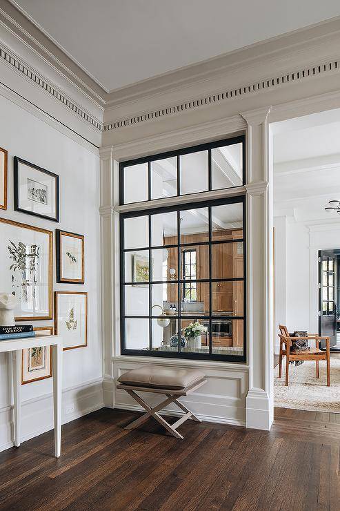 A taupe leather pillow top x-stool sits on a wood floor beneath interior windows, as walls are lined with dentil crown molding.