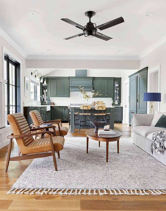 A black ceiling fan cools an open living room featuring an oval wooden coffee table placed on a gray fringe rug and flanked by a pebble gray sofa topped with an emerald green velvet pillow and mid-century modern leather and wooden chairs.