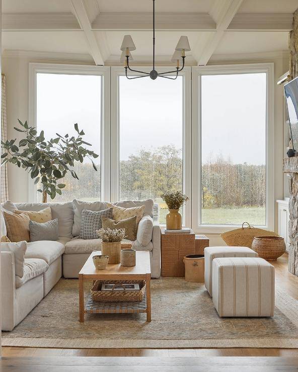 Beneath a coffered ceiling fitted with an oil rubbed bronze chandelier, a welcoming family room features a pebble gray sectional complemented with yellow and brown pillows and placed on layered rugs beside woven nesting end tables. Beige striped cube stools are positioned in front of a brown wooden coffee table, as natural light streams in from tall picture windows.