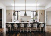 A row of black and white upholstered stools are placed in a brown kitchen at a light brown chevron island lit by gold and black conical pendants hung over a trough sink with