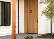 A white painted brick home features a pergola that sits over a plank front door with a bronze ring door knocker.