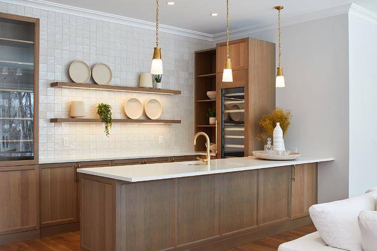 Lighted oak floating shelves are stacked against light gray grid backsplash tiles over oak wet bar cabinets adorned with brass hardware. The shelves are flanked by glass front cabinets and brown oak shelves, while white and gold mini lanterns hang over a brown oak island finished with a sink and a brass gooseneck faucet.