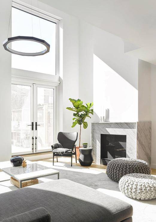 Chic modern living room boasts gray knitted stools placed on a gray rug in front of a modern gray marble fireplace, as a black accent table is paired with a black leather chair located in front of a fiddle leaf fig.