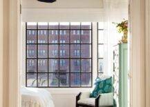 Bedroom with transom window inviting natural light highlighting vintage style decor and furnishings. A black and white chair is furnished in a corner beside a green tall boy vintage dresser with ring pulls finished with a pink and blue area rug under a bed.