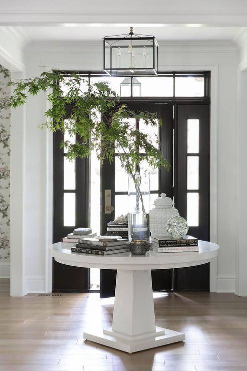 Flanked by sidelights and positioned under a transom window, a black front door with glass panels opens to a foyer boasting a round white styled entry table lit by a flush mount lantern.