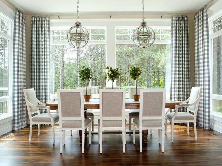 Chic dining room features a trestle dining table lined with heather gray square back French dining chairs illuminated by Restoration Hardware Victorian Hotel Pendants surrounded by windows dressed in blue gingham curtains.