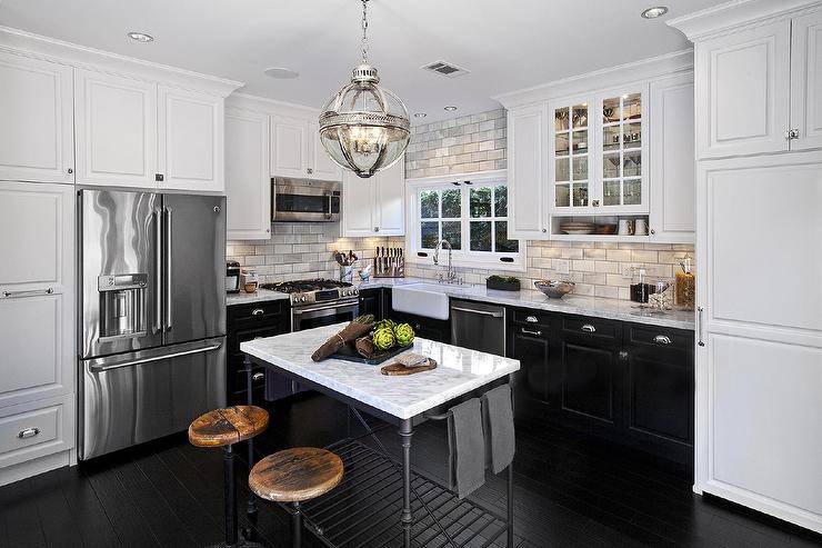 L shaped kitchen features white upper cabinets and black lower cabinets paired with marble countertops and a marble subway tiled backsplash. A small farmhouse sink and a vintage gooseneck faucet stands adjacent to a microwave hood and stainless steel stove. A Restoration Hardware Victorian Hotel Pendant illuminates an iron and marble kitchen island, Crate & Barrel French Kitchen Island, lined with two industrial swivel bar stools placed atop an ebony floor.