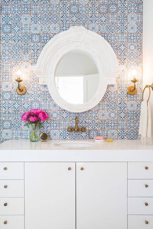 White and blue bathroom features a wall clad in blue Moroccan tiles, Fez Blue Vintage Moroccan Victorian Encaustic Effect Pattern Wall & Floor Tiles, lined with a white lacquered washstand adorned with aged brass knobs topped with thick white quartz and paired with an aged brass faucet placed under a white Mansard mirror.