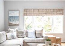 Light beige sectional in a living room paired with a light brown leather ottoman styled with stacked wood trays and a floral centerpiece. Various gray and white accent pillow style the sectional with a contemporary add on while a set of woven nesting end tables deliver a warm Scandinavian like feel.