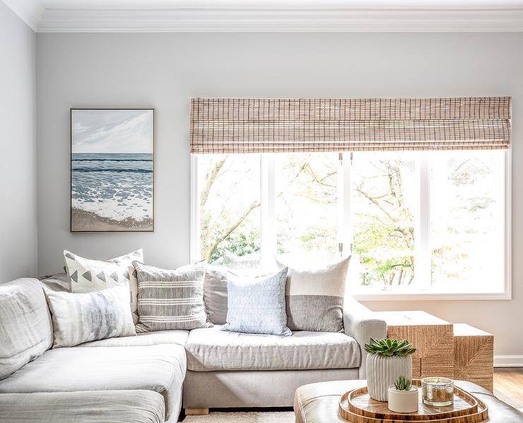 Light beige sectional in a living room paired with a light brown leather ottoman styled with stacked wood trays and a floral centerpiece. Various gray and white accent pillow style the sectional with a contemporary add on while a set of woven nesting end tables deliver a warm Scandinavian like feel.