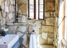 Small rustic cottage bathroom features a vanity mirror mounted to a stone covered wall beside a Reed Single Sconce and over a black washstand topped with a white overmount sink fitted with an oil rubbed bronze faucet. Rustic wood floor paneled covered in a blue and red rug leads to a walk in shower separated by a stone partition and fitted with tumbled marble brick floor tiles, a stone surround, and an oil rubbed bronze shower kit lit by natural light from a casement window.