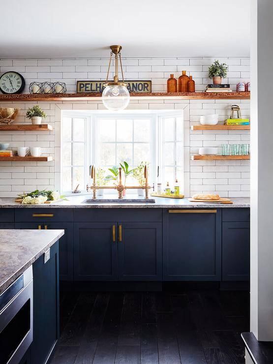 Brown wooden floating shelves are fixed to white subway wall tiles on either side of and above a window located above a sink with a brass dual deck mount faucet. The faucet is mounted to a gray granite countertop finishing blue shaker cabinets adorned with brass hardware and placed on an ebony wood floor.