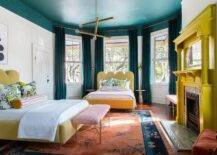 Contemporary bedroom features a yellow painted fireplace mantel with mosaic tiles, a yellow scalloped headboard with lemon print pillows and a pink stripe bolster pillows under a bay window with peacock blue velvet curtains, a matching bed under art and a peacock blue painted ceiling.