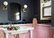 Gold and black sconces are mounted to a black painted wall on either side of a curved vanity mirror hung above pink offset wall tiles finished with black border tiles. A white French sink vanity is fixed in front of the backsplash tiles and against black and white hexagon floor tiles.