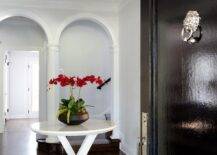 Crystal cascading chandelier illuminates a foyer furnished with a small round white trestle accent table on a dark gray rug with a white border. A glossy black front door with a silver lion door knocker brings a classic appeal to the entryway of the home.