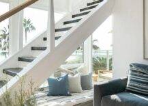 A small reading nook is located beneath a staircase and is accented with a rectangular gray floor cushion topped with a thin blue striped cushion and white and blue pillows.