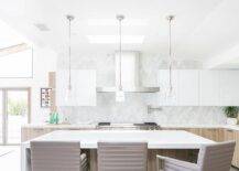 Taupe leather counter stools sit at a brown oak kitchen island topped with a white quartz waterfall countertop lit by mini rippled glass island lights and natural light from a skylight.