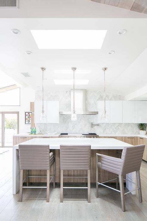 Taupe leather counter stools sit at a brown oak kitchen island topped with a white quartz waterfall countertop lit by mini rippled glass island lights and natural light from a skylight.