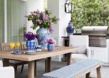 Brown teak dining table and a matching dining bench feature a blue and white pattern upholstered cushion. Natural linen slipcovered dining chairs mix with the bench and teak chairs atop slate pavers next to a covered patio bbq.