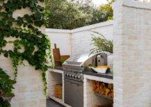 Charming outdoor kitchen nook boasts off-white brick walls, slate pavers, and an stainless steel BBQ flanked by a black countertop located over a firewood storage shelf.