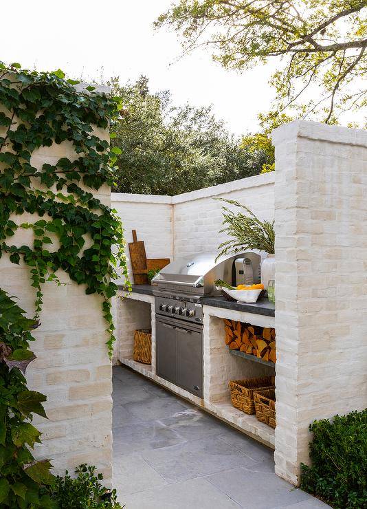 Charming outdoor kitchen nook boasts off-white brick walls, slate pavers, and an stainless steel BBQ flanked by a black countertop located over a firewood storage shelf.