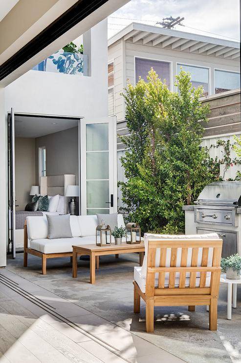 A teak chair and a teak sofa accented with white cushions and gray pillows sit on either side of a teak coffee table positioned to the side of a BBQ.