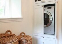 Dream walk-in closet features washer dryer combo hidden within built-in white cabinets with glass front uppers, accented with mercury glass pulls alongside a pair of seagrass laundry hampers.