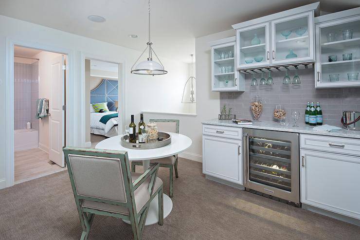 Ikea Docksta Table in a second-floor landing kitchenette is paired with green and gray distressed dining chairs surround white and gray decor. Gray subway tiles create a neutral backsplash around upper cabinets with frosted glass front doors displaying light blue kitchenware. An under counter wine cooler is flanked by white cabinets with steel hardware under marble countertops presenting a luxurious touch to the landing kitchen design.