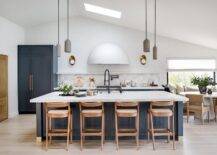 Staggered gray pendants hang from a vaulted ceiling fitted with a skylight on either side of a blue kitchen island. The island holds a sink with an oil rubbed bronze gooseneck faucet in front of brown wooden counter stools.