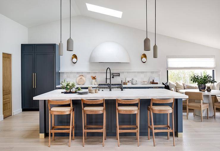 Staggered gray pendants hang from a vaulted ceiling fitted with a skylight on either side of a blue kitchen island. The island holds a sink with an oil rubbed bronze gooseneck faucet in front of brown wooden counter stools.