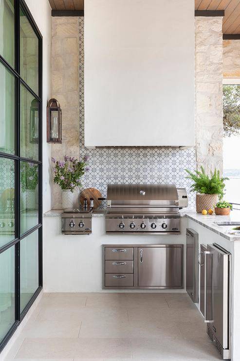 Well appointed l-shaped outdoor kitchen boasts a white hood fixed to gray and blue mosaic concrete backsplash tiles over a stainless steel BBQ.