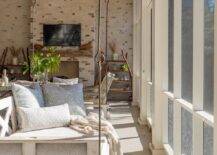 Welcoming sunroom features a whitewashed wooden swing sofa topped with pale blue pillows and hung from a brown wood plank ceiling.