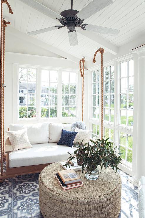 Hung in a gorgeous sunroom from a vaulted plank ceiling, a wood and rope swing sofa is accented with white cushions and hangs over gray quatrefoil floor tiles facing a round woven ottoman.