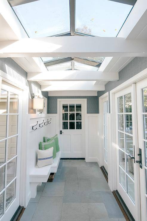 A vaulted ceiling fitted with skylights allows natural light to stream into to a sunroom boasting slate floor tiles and a white built-in bench mounted beneath a row of iron hooks fixed to a shiplap backsplash lined with a gray upper wall.