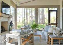 Welcoming gray sunroom features a gray wicker and teak couch placed on a gray rug beside a teak and concrete end table and facing a teak and concrete coffee table. The coffee table is flanked by gray wicker and teak accent chairs, while the space is completed with a white painted brick fireplace finished with a rustic wood mantel and wall mounted TV.